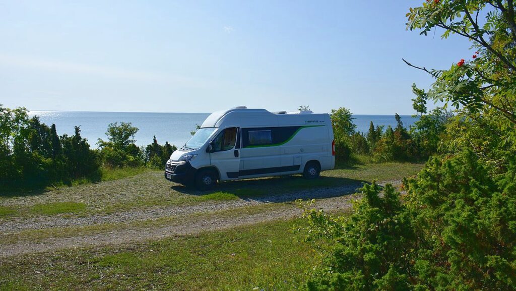 En liten husbil parkerad på grus, omgiven av natur och med hav i bakgrunden