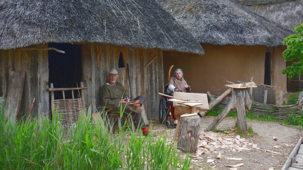 Kaksi henkilöä viikinkiaikaisissa vaatteissa viikinkiaikaisten rakennusten edustalla