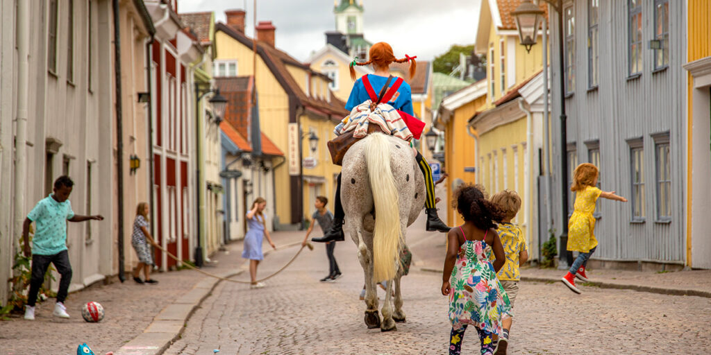 Pippi Långstrump rider på gatorna i en stad och barn leker runt omkring